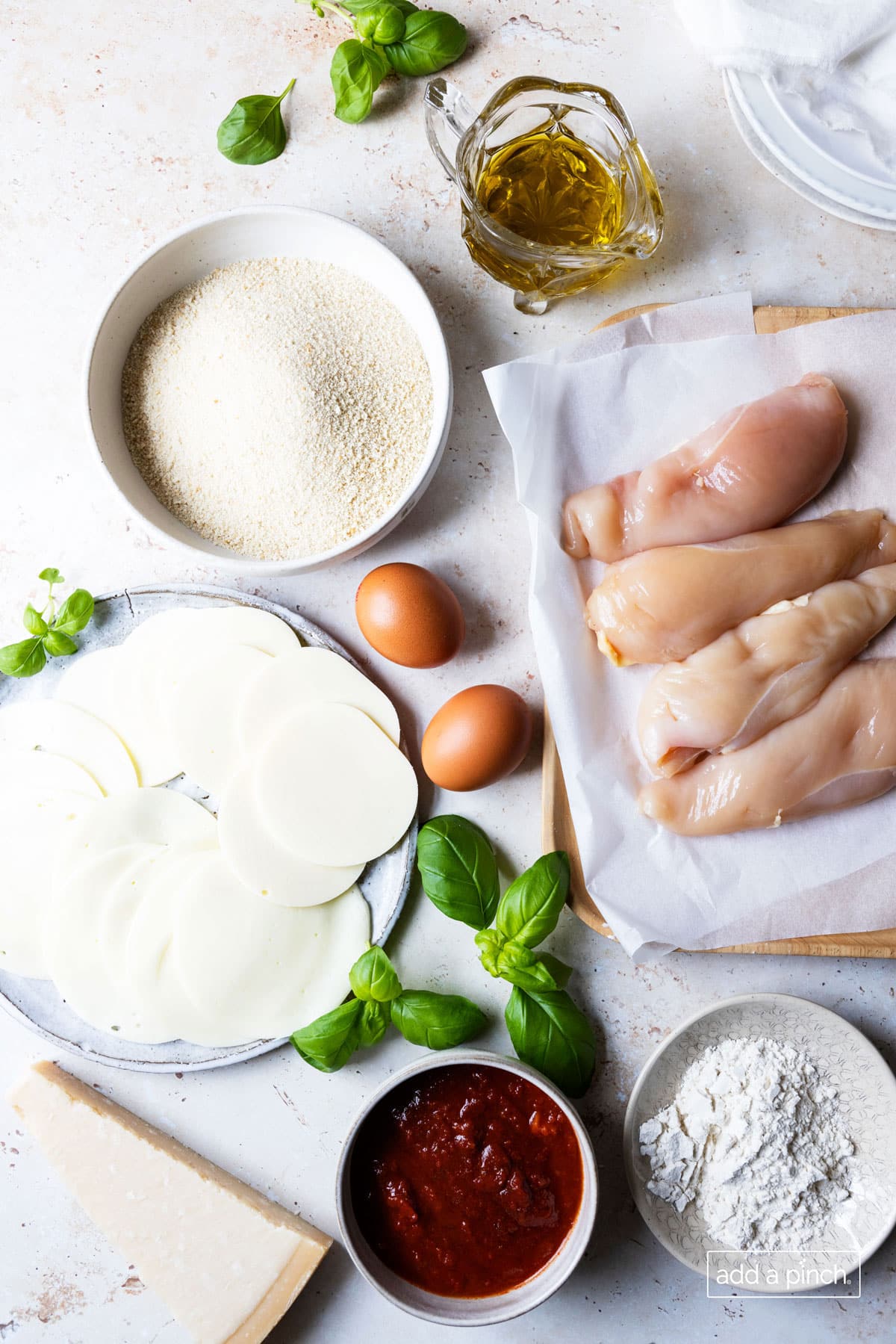 Photograph of ingredients needed for chicken parmesan: chicken breasts, eggs, flour, marinara sauce, mozzarella cheese slices and grated parmesan cheese.