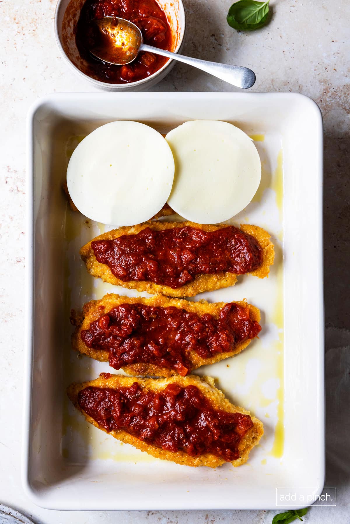 Photo of browned chicken in a white baking dish topped with marinara sauce and cheese.