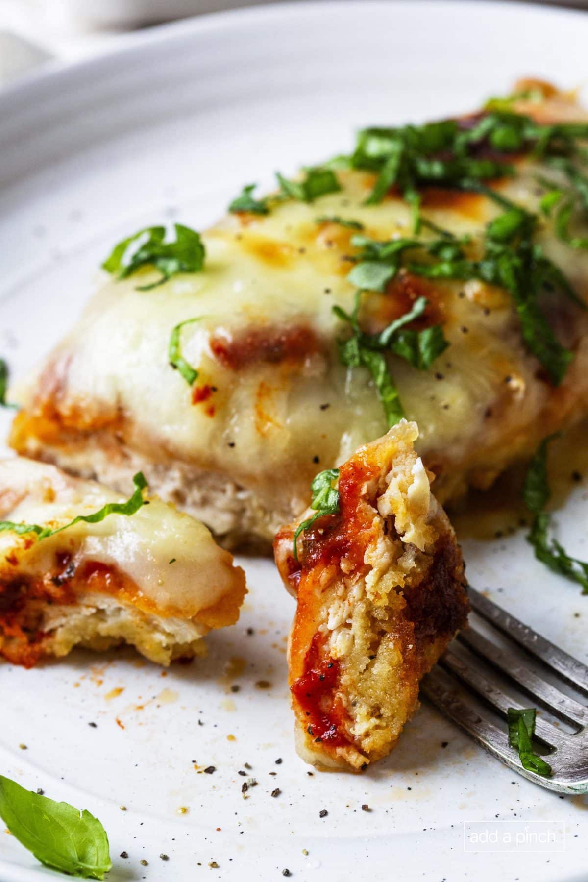 Photograph of chicken parmesan recipe with a bite on a fork served on a white plate.