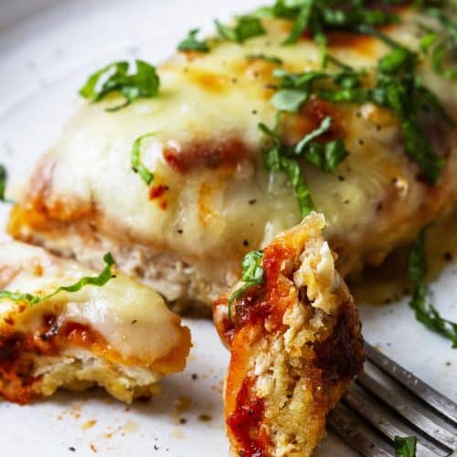 Photograph of chicken parmesan recipe with a bite on a fork served on a white plate.