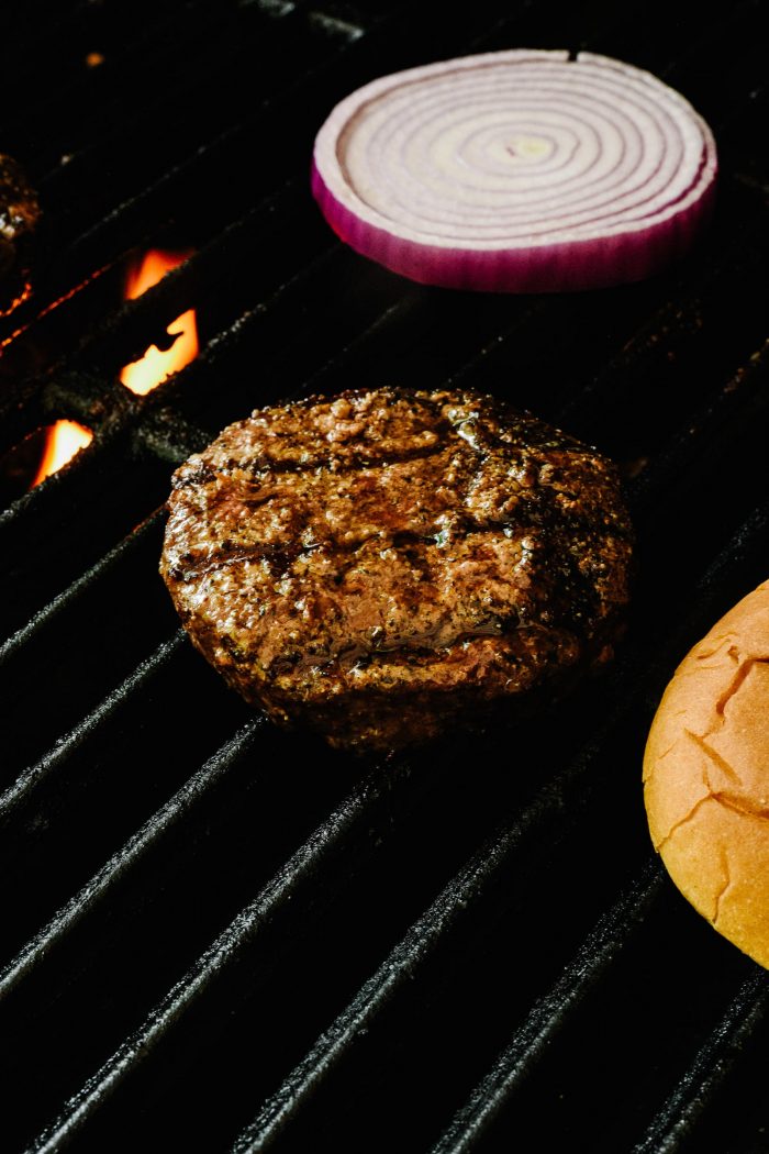 Grill grate with flame under it, with juicy grilled burger, onion slice and bun on grate // addapinch.com