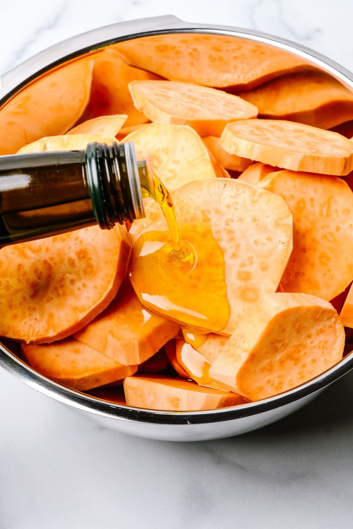 Olive oil being poured over slices of raw sweet potatoes in stainless bowl. // addapinch.com 