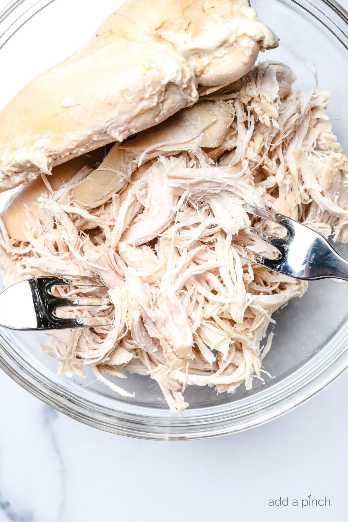 Photograph of chicken in a glass bowl being shredded with two forks.