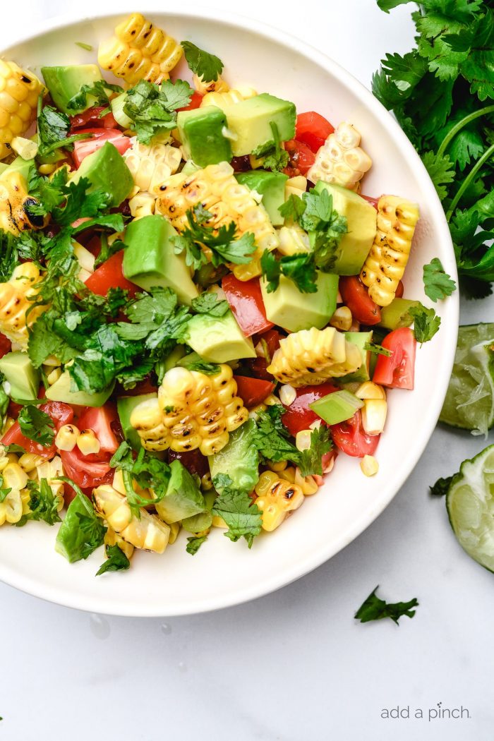 Grilled corn, avocado, diced tomatoes, chopped cilantro and green onion in a bowl surrounded by fresh cilantro and lime. // addapinch.com