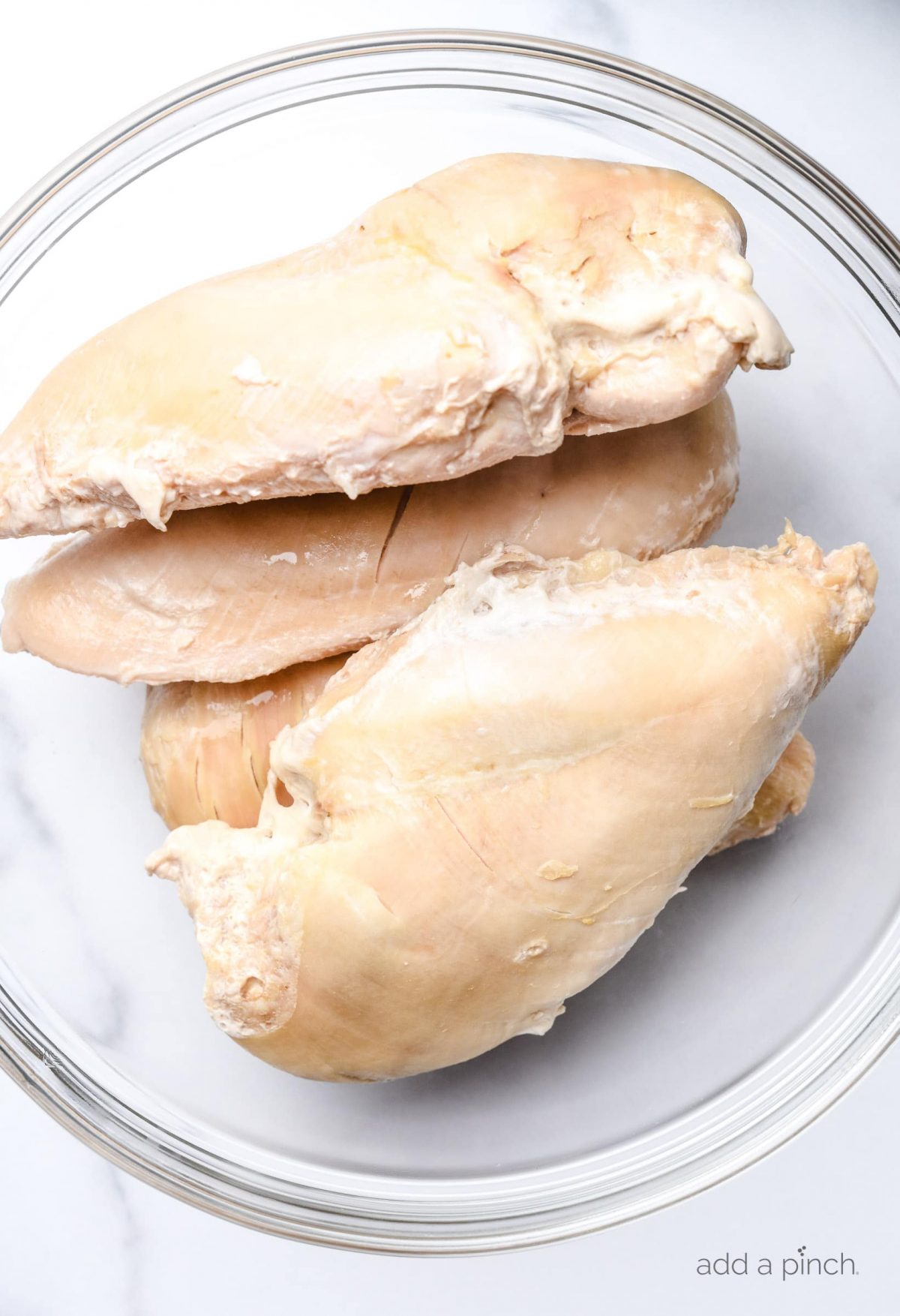 Four cooked chicken breasts in a clear glass bowl on a white marble surface.