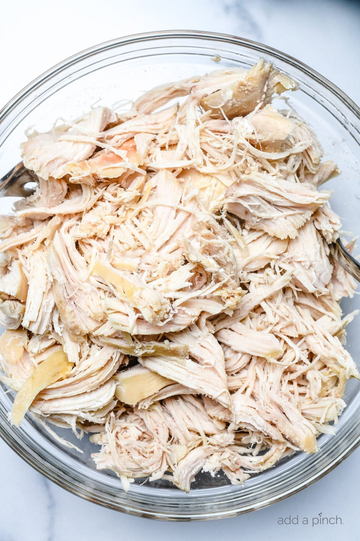 Cooked and shredded chicken in a glass bowl on a marble surface.