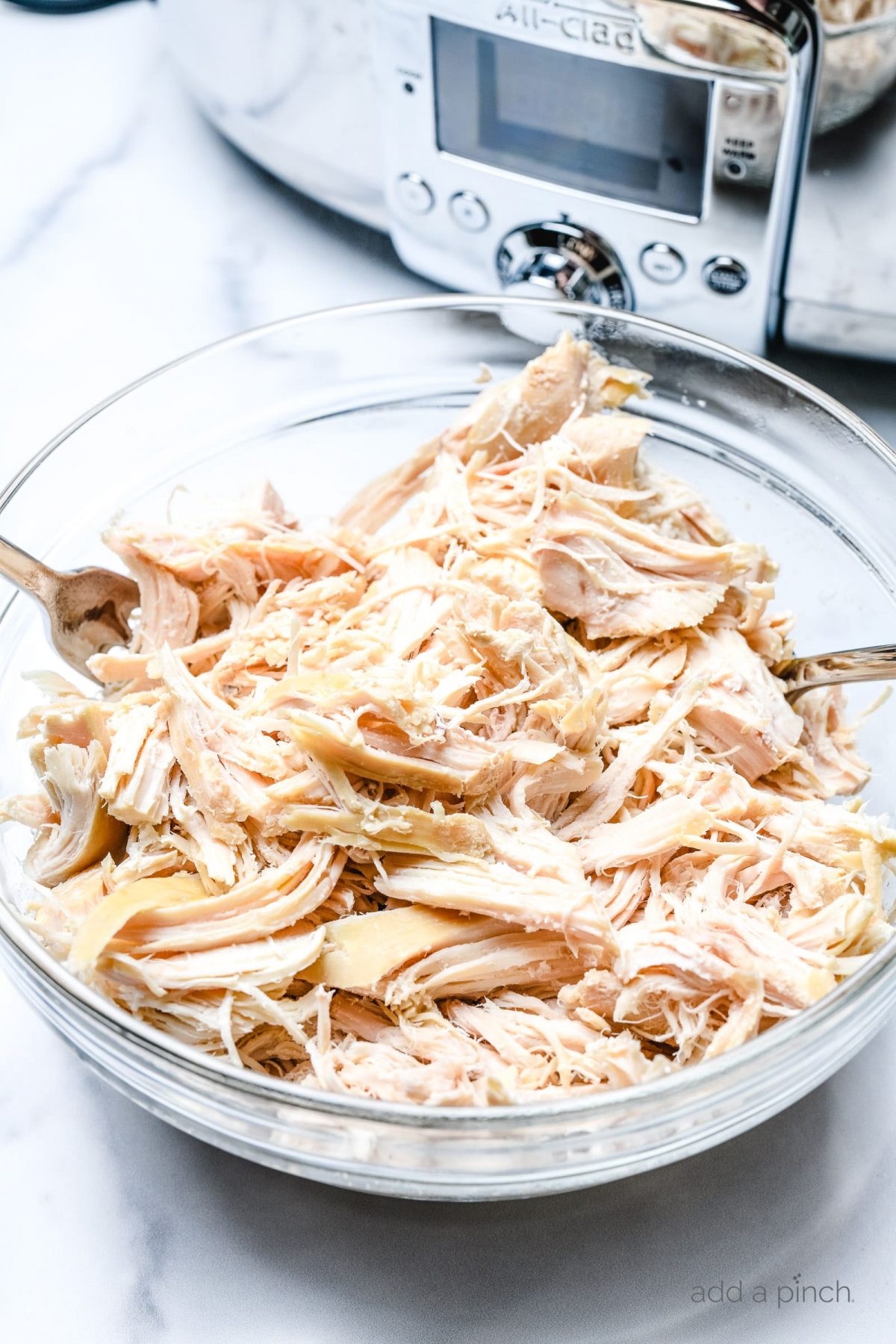 Shredded chicken in a clear glass bowl with two forks in the bowl and an instant pot in the background.