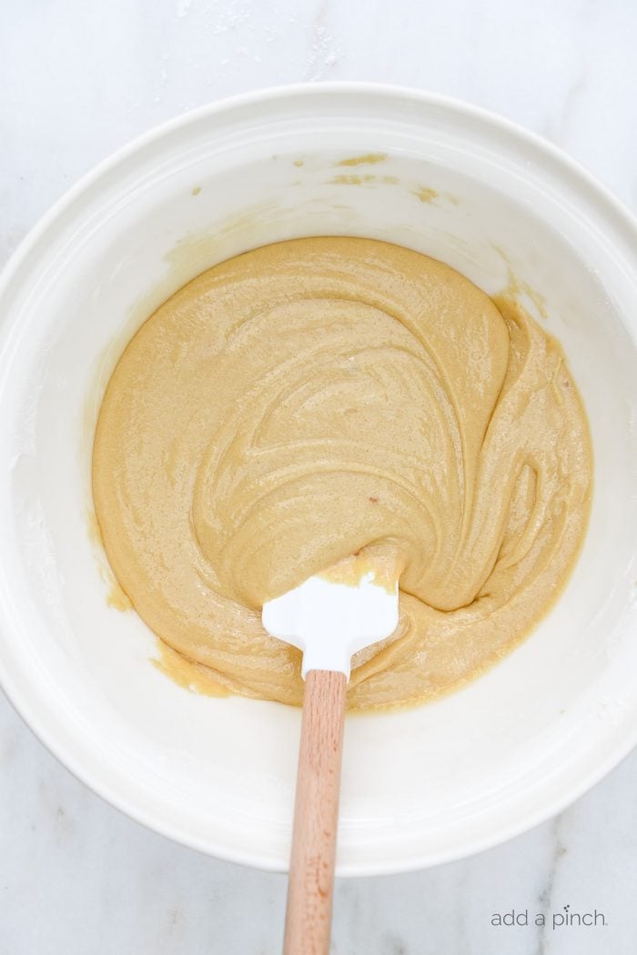 White mixing bowl and wooden handled spatula with blended batter for Pecan Chewies. // addapinch.com