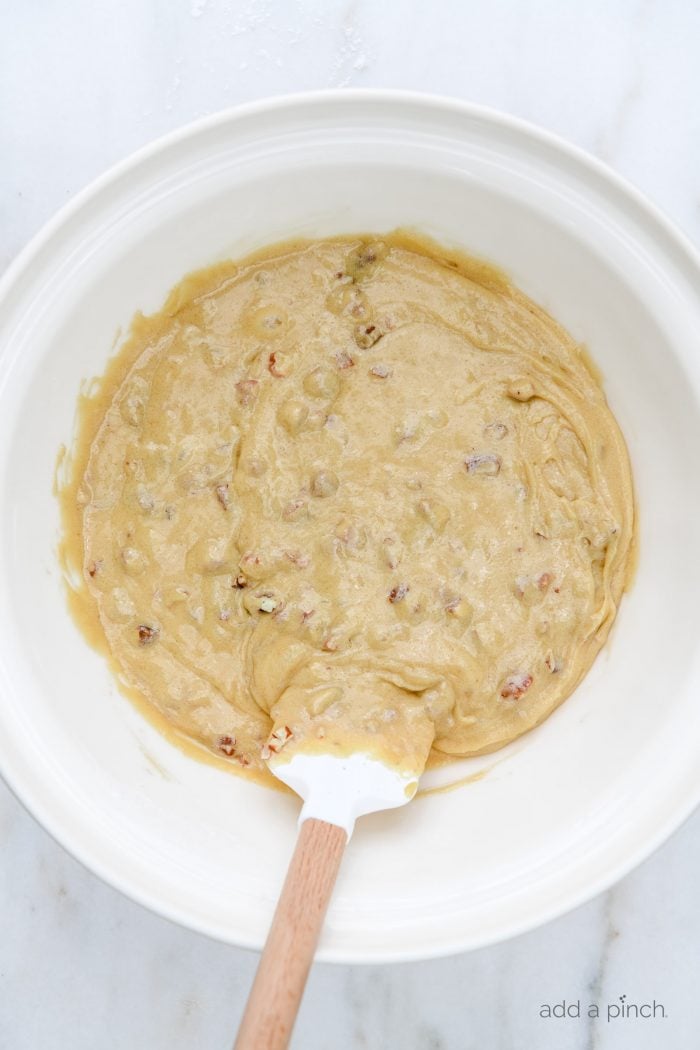 Chopped pecan are blended into batter with wooden handled spatula into white mixing bowl. // addapinch.com