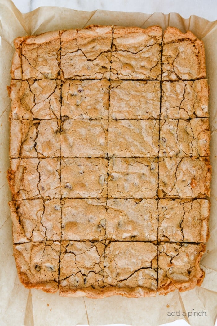 Brown parchment paper lifted from baking pan, with golden baked Pecan Chewies cut into squares.  