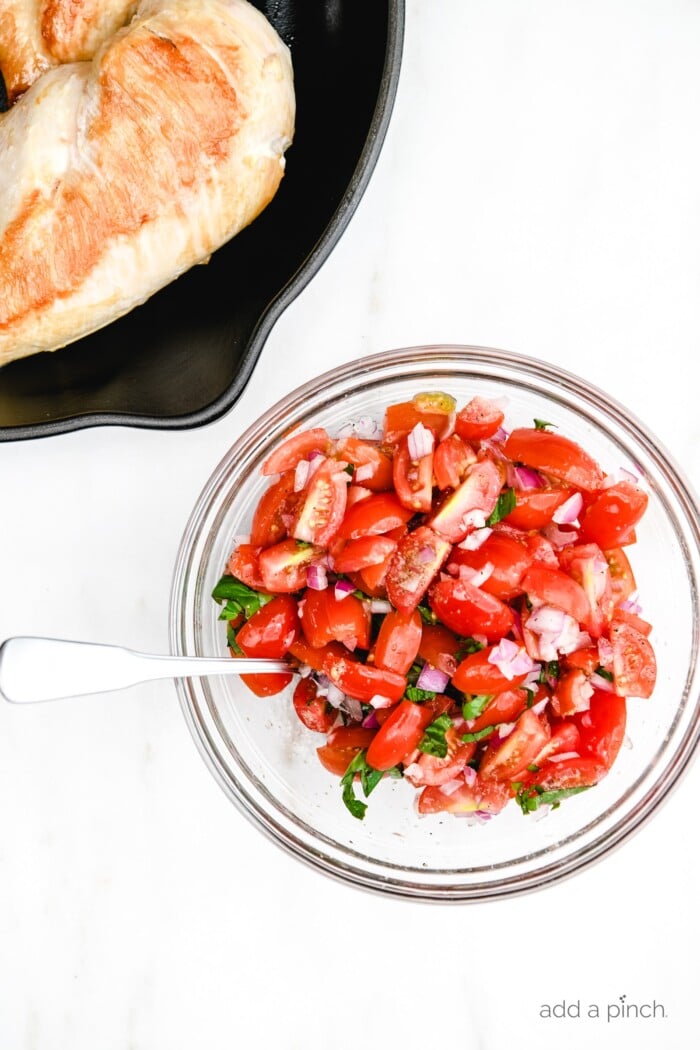 Chicken breast in a skillet and bruschetta mixture in a glass bowl with a spoon.