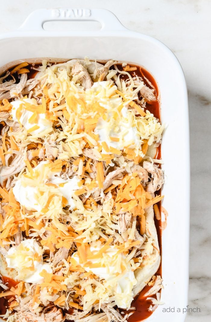 Layered ingredients of chicken, tortillas, cheese and sour cream on top of enchilada sauce // addapinch.com