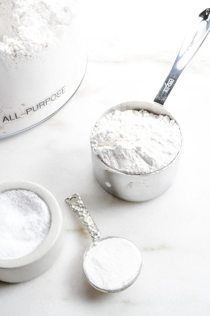 Ingredients to make a substitute for self-rising flour sit on a marble countertop. Salt, baking powder and all-purpose flour are all measured for mixing.