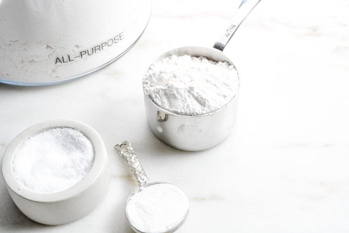 Measuring cup of flour, salt and measuring spoon of baking powder sit on marble counter beside a glass container of flour.