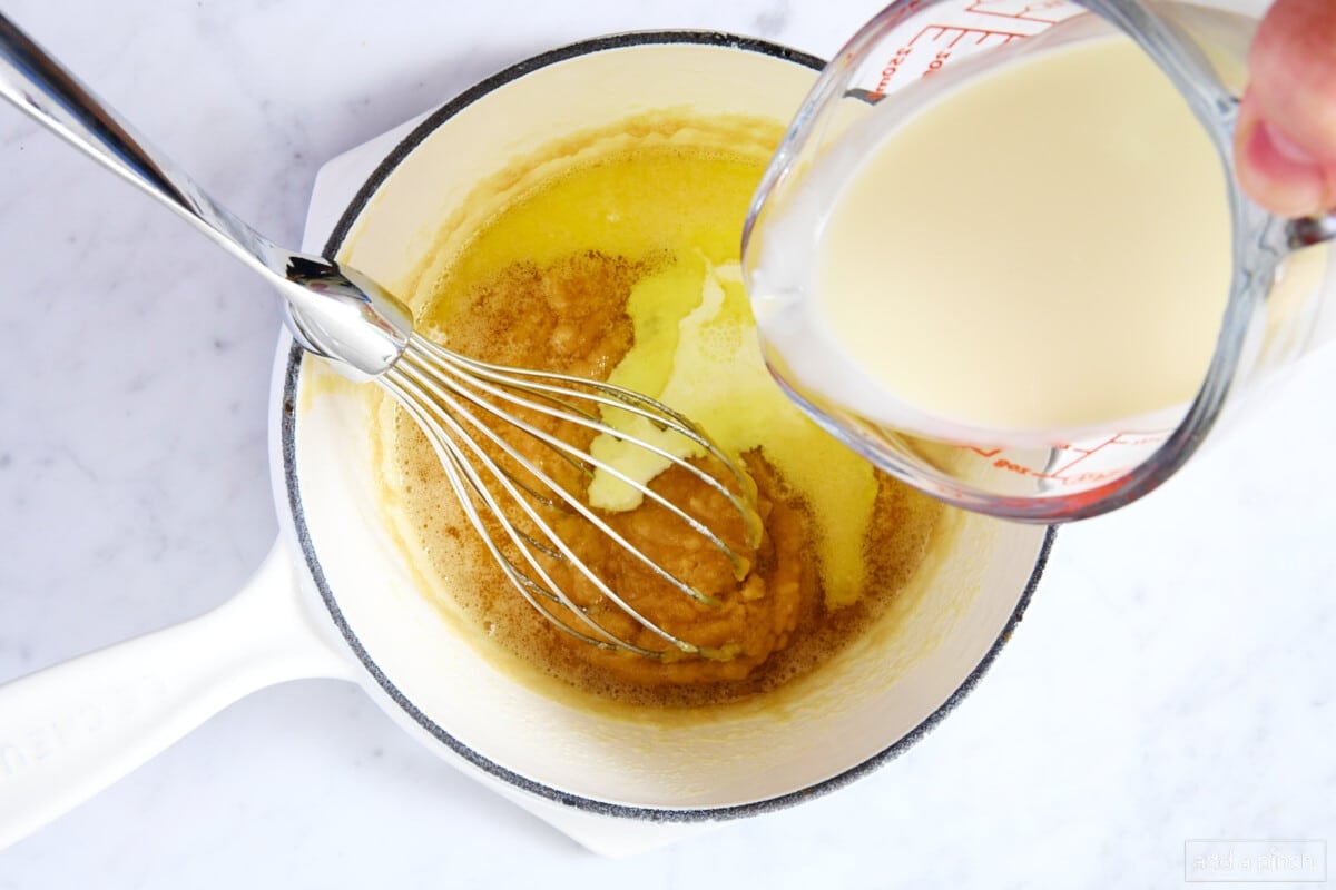Pouring heavy cream to caramel sauce in a white saucepan with a metal whisk.