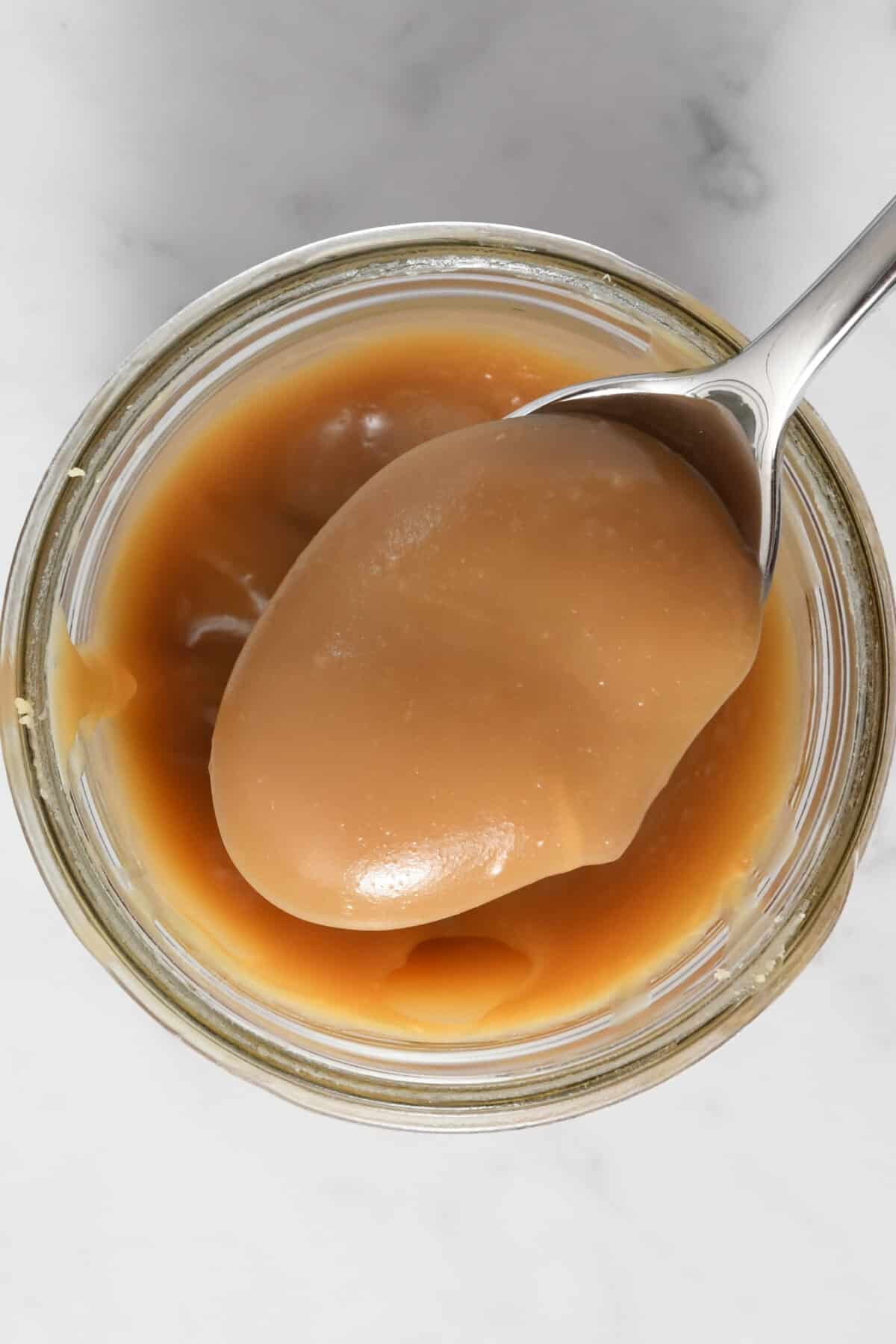 A metal spoon with a scoop of caramel sauce in a glass jar.