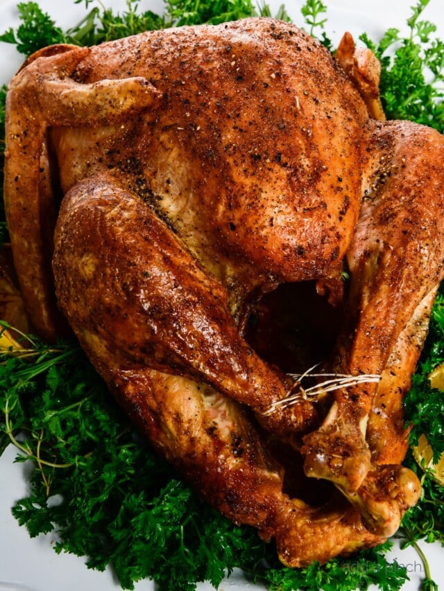 Photograph of golden brown roasted turkey served on a white platter with fresh herbs.