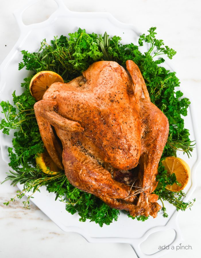 Overhead photo of a roasted turkey on a large white platter garnished with fresh herbs, lemon, salt and pepper. // addapinch.com