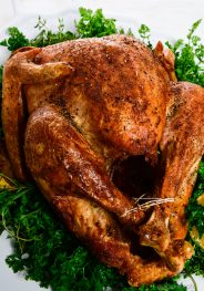 Photograph of golden brown roasted turkey served on a white platter with fresh herbs.