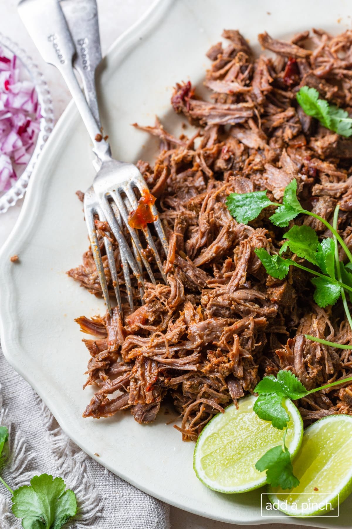 Barbacoa on a platter with cilantro, lime slices, and two forks. 