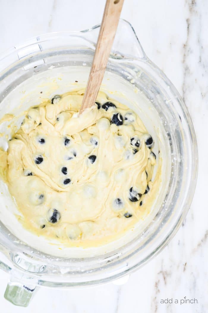 Blueberry muffin batter in a glass bowl with a wooden spoon on marble countertop.