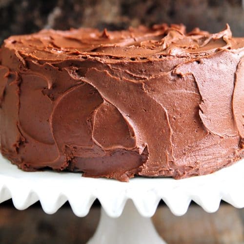 Chocolate Cake with Chocolate Buttercream Frosting on a white cake plate and dark background.
