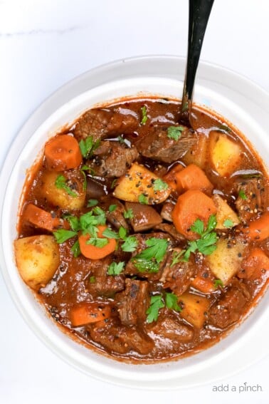 Picture of beef stew with carrots, potato chunks, tomato sauce, parsley, chunks of beef in a white bowl.