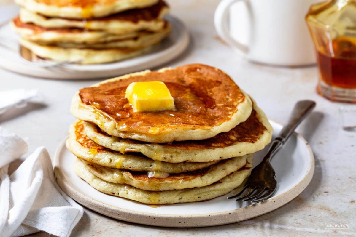 Table set with plates filled with stacks of pancakes topped with butter and syrup. 
