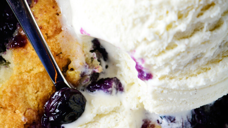 Photo of blueberry cobbler topped with vanilla ice cream in white bowl.