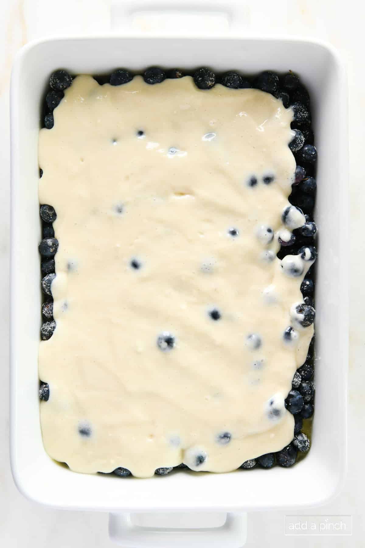 Cobbler batter poured over sugared blueberries in a white baking dish.