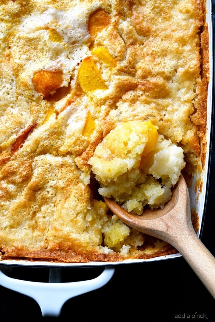 Overhead photograph of spoonful of easy peach cobbler in a white baking dish on a stone counter. // addapinch.com