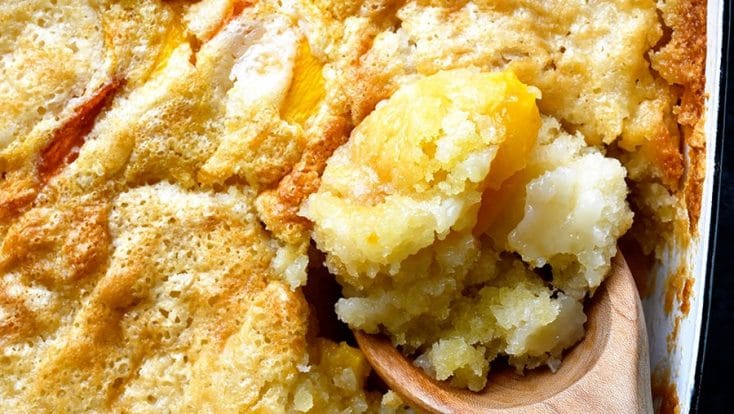 Overhead photograph of spoonful of easy peach cobbler in a white baking dish on a stone counter. // addapinch.com