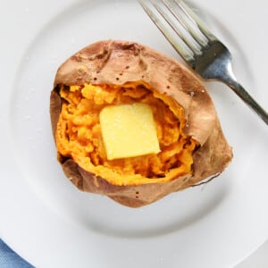 photo of baked sweet potato topped with butter and salt on a white plate with a fork.