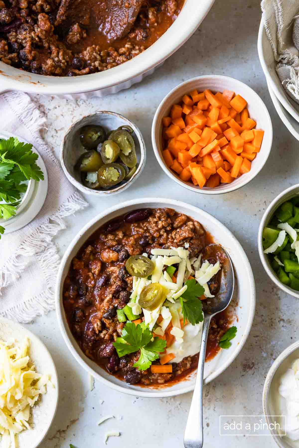 Chili topped with cheese, peppers, and fresh herbs, surrounded by bowls of optional toppings.