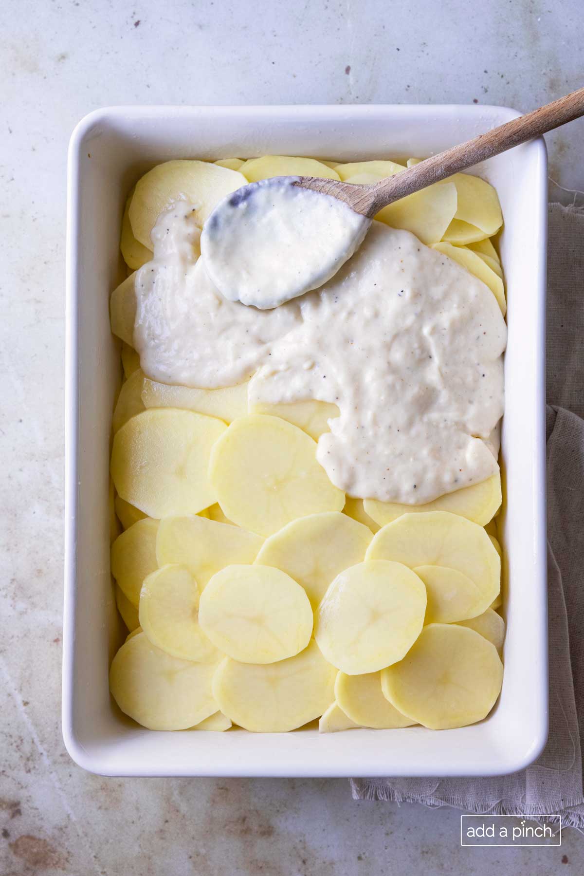 Photo of sliced potatoes in a white baking dish with cream sauce being spread over the top with a wooden spoon.