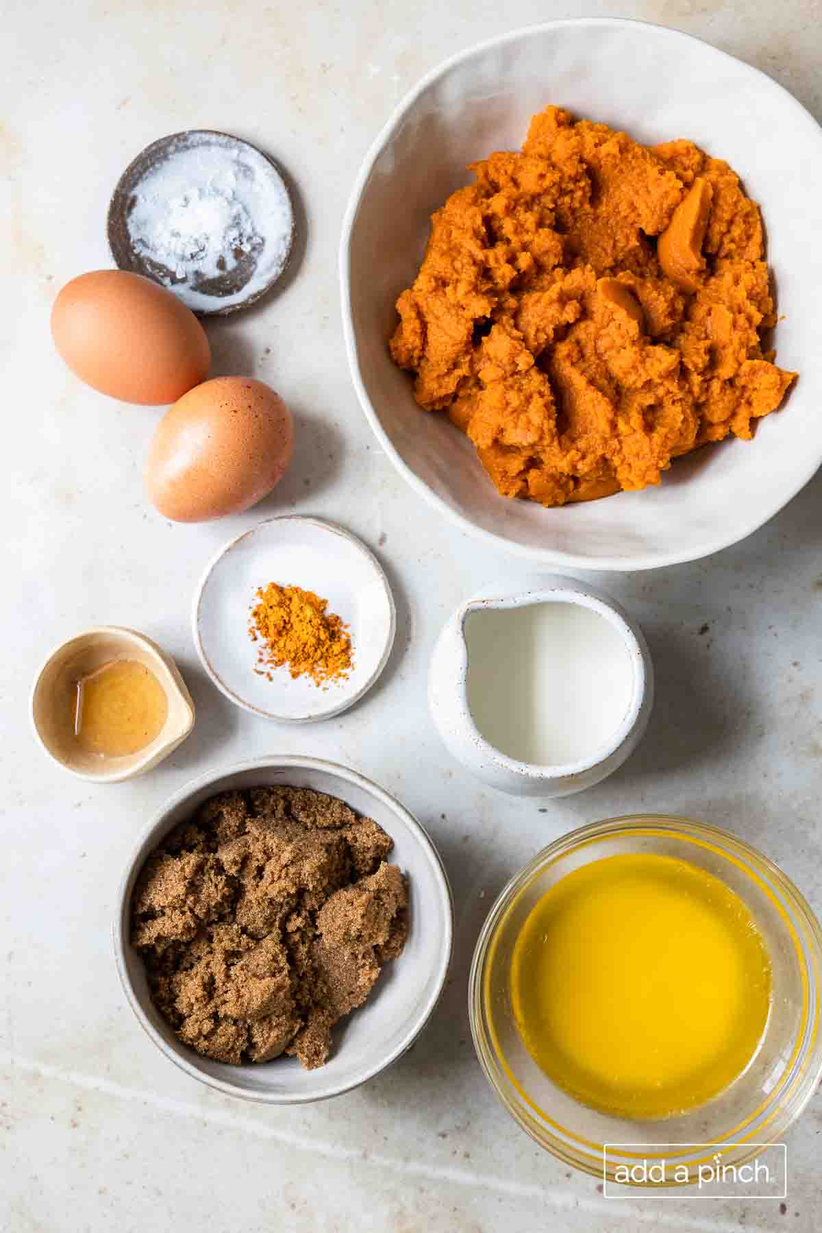 Pumpkin pie ingredients like melted butter, brown sugar, half and half, spices, eggs, salt and pumpkin puree all on a white stone counter.