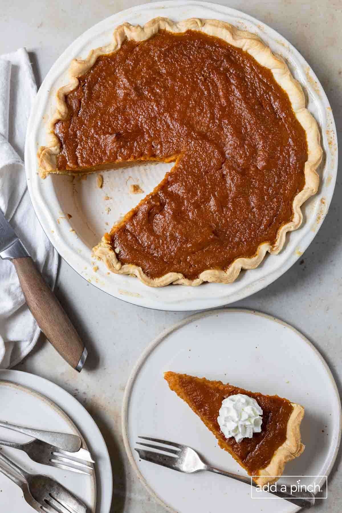 White pie plate holds a pumpkin pie with slice cut and served on a plate with fork and whipped cream on top.