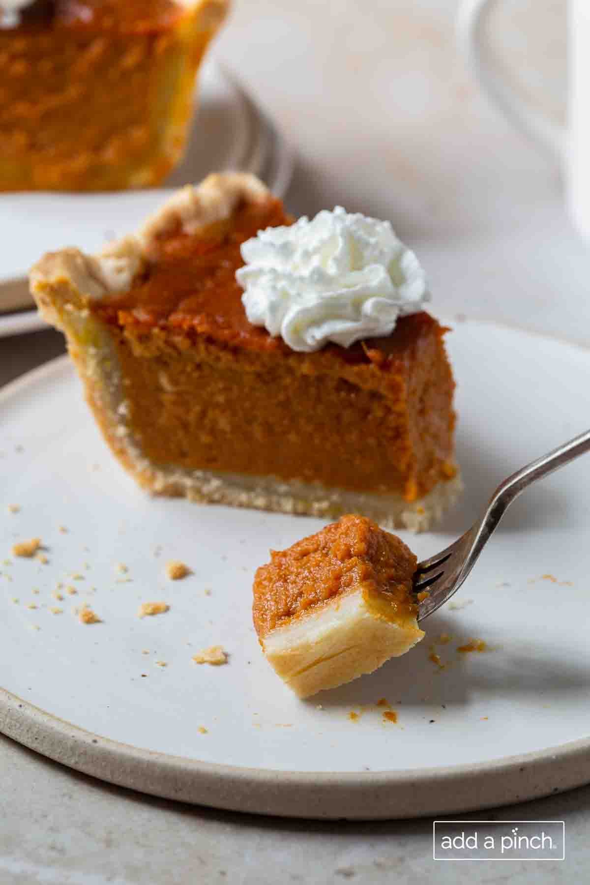 Slice of pumpkin pie with whipped cream on top with a bite cut onto a fork, surrounded by scattered pie crust crumbs, on a white plate - addapinch.com