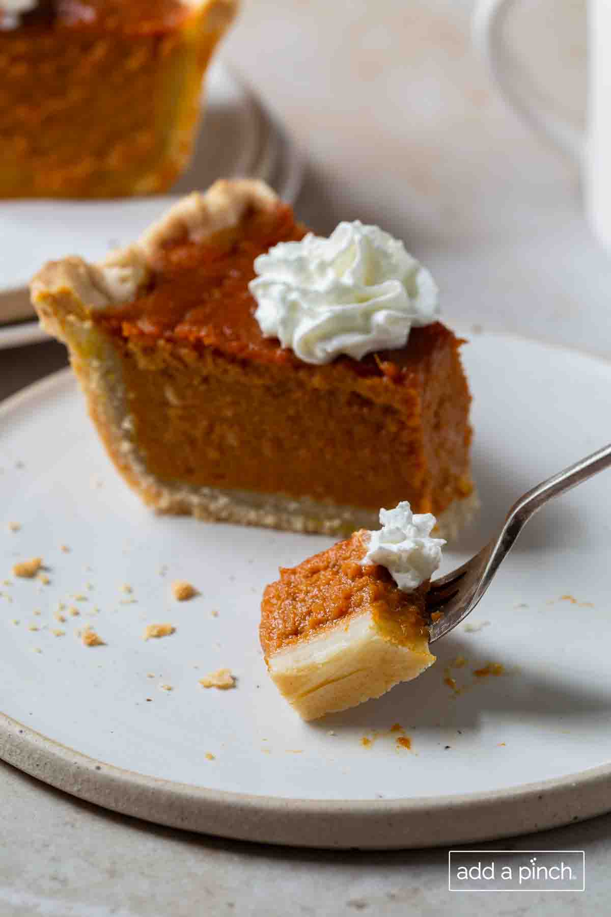 Slice of pumpkin pie on white plate with a bite on a fork.
