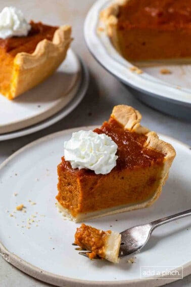Slice of pumpkin pie with one bite on a fork. Remaining pie in the background. All on white surfaces.