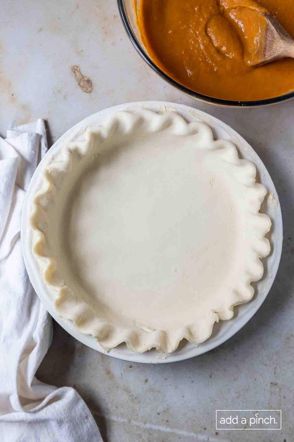 Photo of pie crust dough in a white pie plate with pie filling in a clear glass bowl with a wooden spoon in the background.