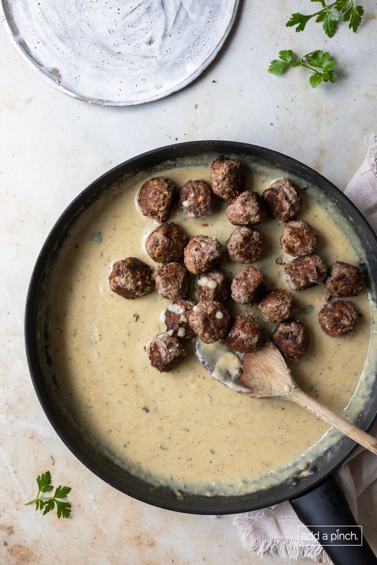 Browned meatballs added to creamy sauce in a skillet with fresh parsley nearby.
