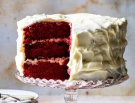 Photograph of red velvet cake with white frosting with a slice cut so that you can see the three layers of the rich, velvety cake.
