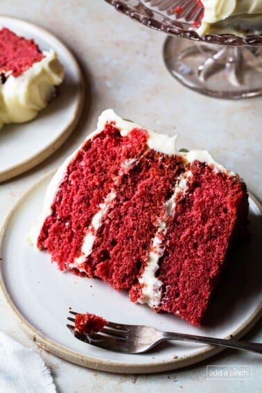 Slice of red velvet cake in three layers frosted with thick cream cheese frosting with slice alongside and cake stand in back.