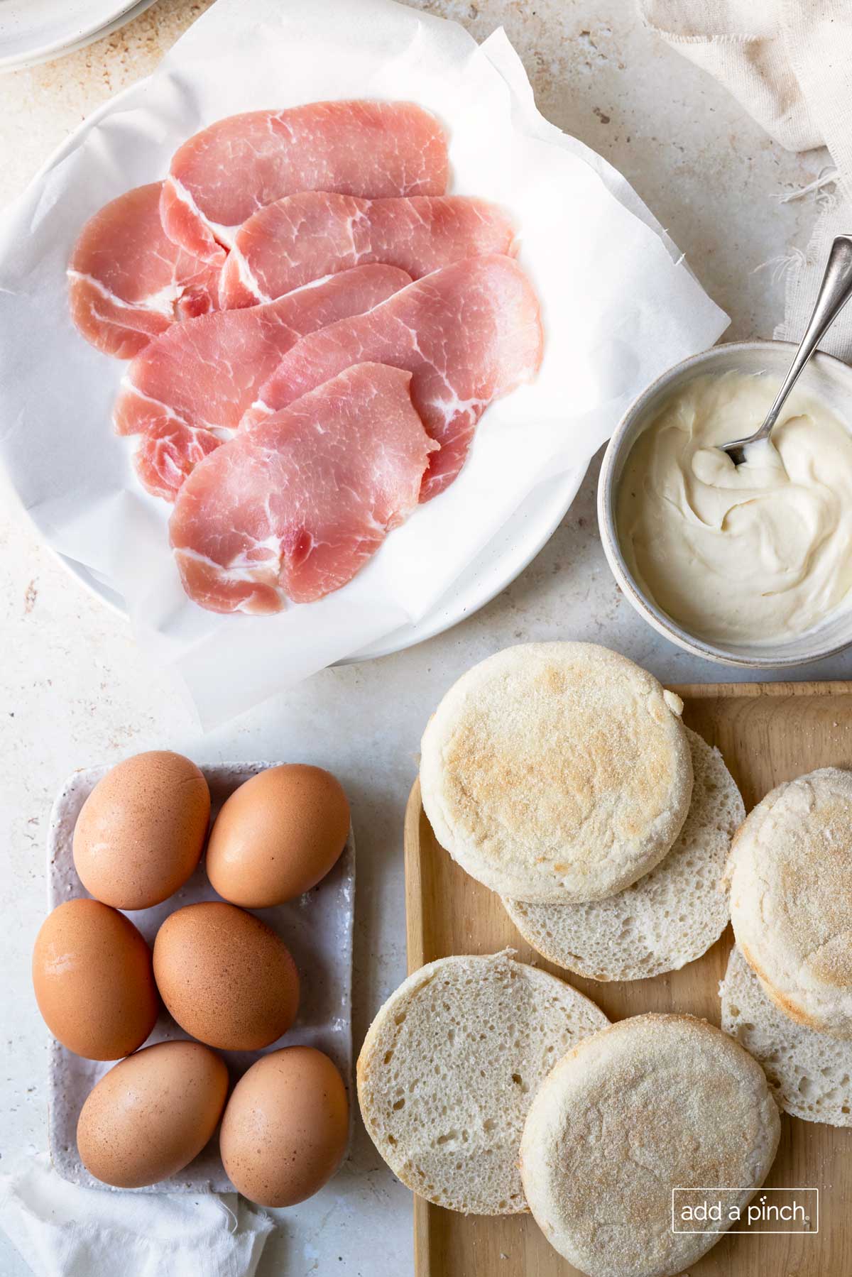 Photo of ingredients needed to make Eggs Benedict: Canadian Bacon, Eggs, English Muffins, Hollandaise Sauce all on a marble surface and wooden cutting board.