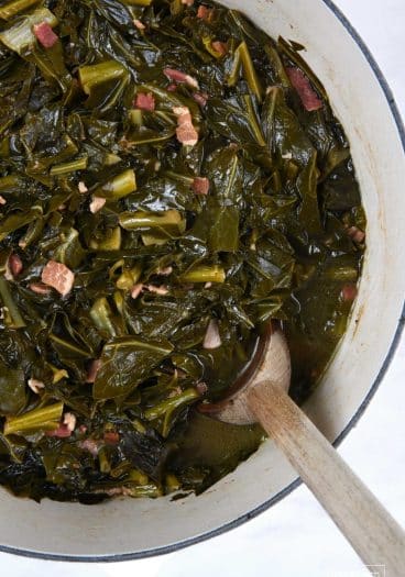 Photo of white pot filled with cooked collard greens and a wooden spoon.