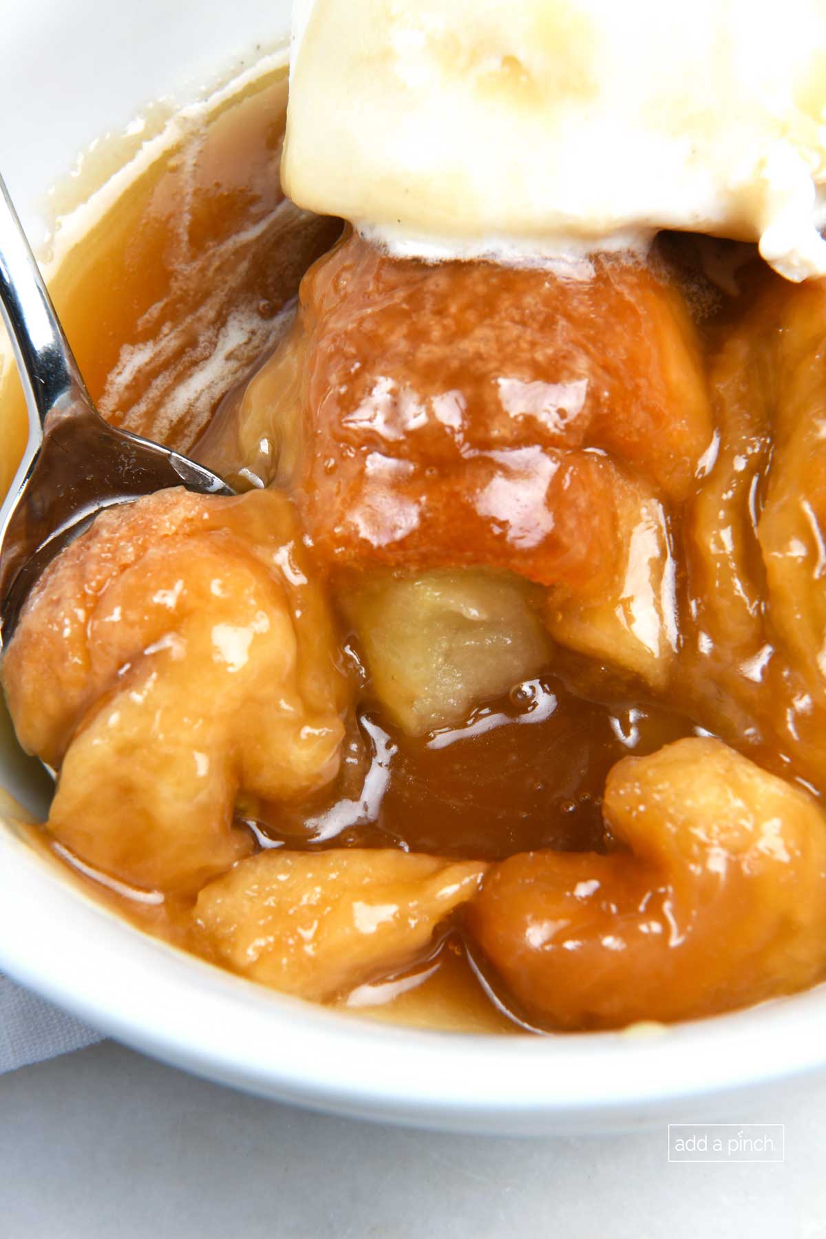 Photo of a spoon filled bite of apple dumpling in a white bowl of dumplings topped with vanilla ice cream and caramel sauce.