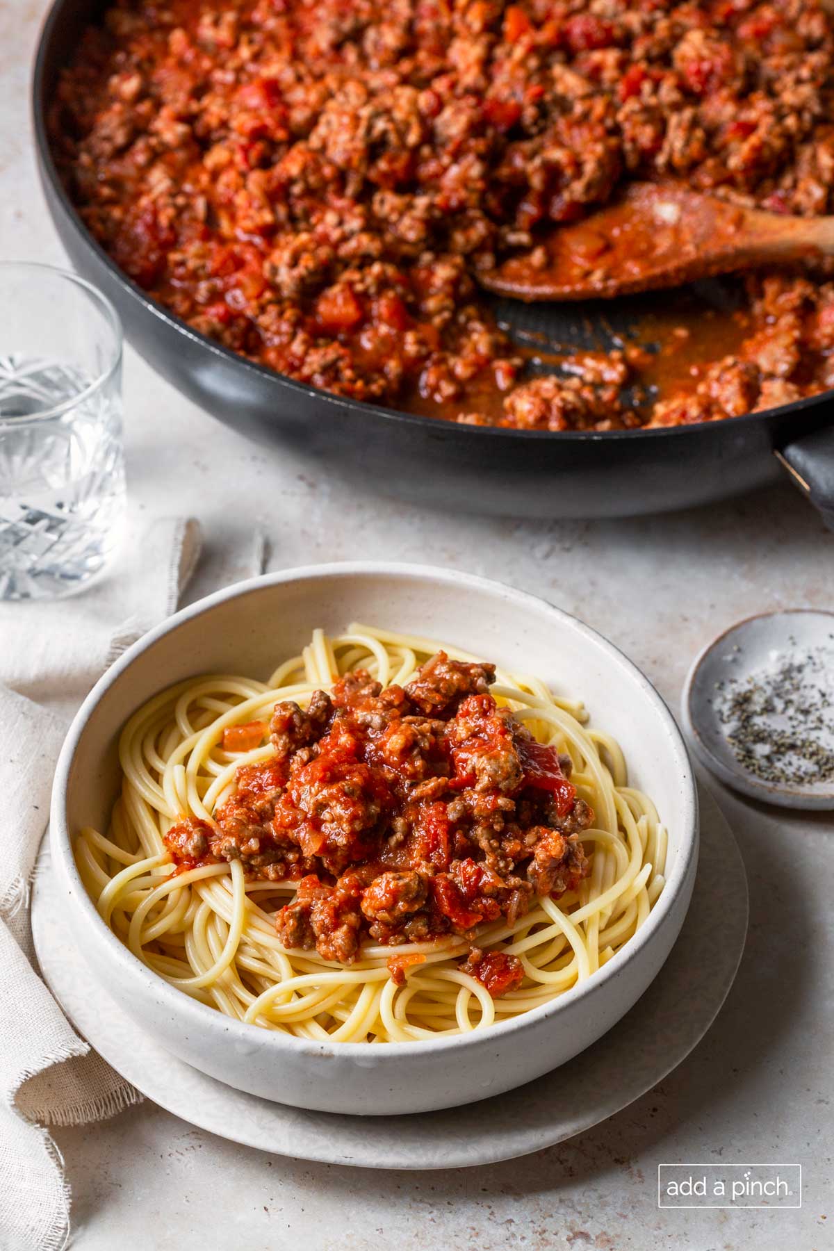 Photo place setting of spaghetti with skillet filled with homemade spaghetti sauce and a wooden spoon in the background.