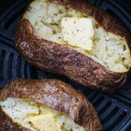 Two air fryer baked potatoes with butter, salt, and pepper in an air fryer basket.