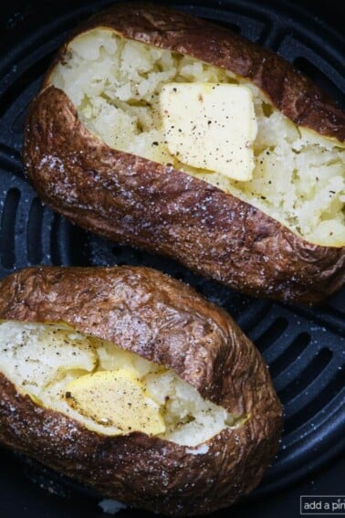 Two air fryer baked potatoes with butter, salt, and pepper in an air fryer basket.